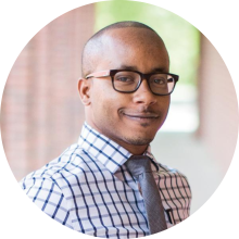 Headshot of Reggie wearing a plaid shirt, tie and glasses standing in the Rotunda. 