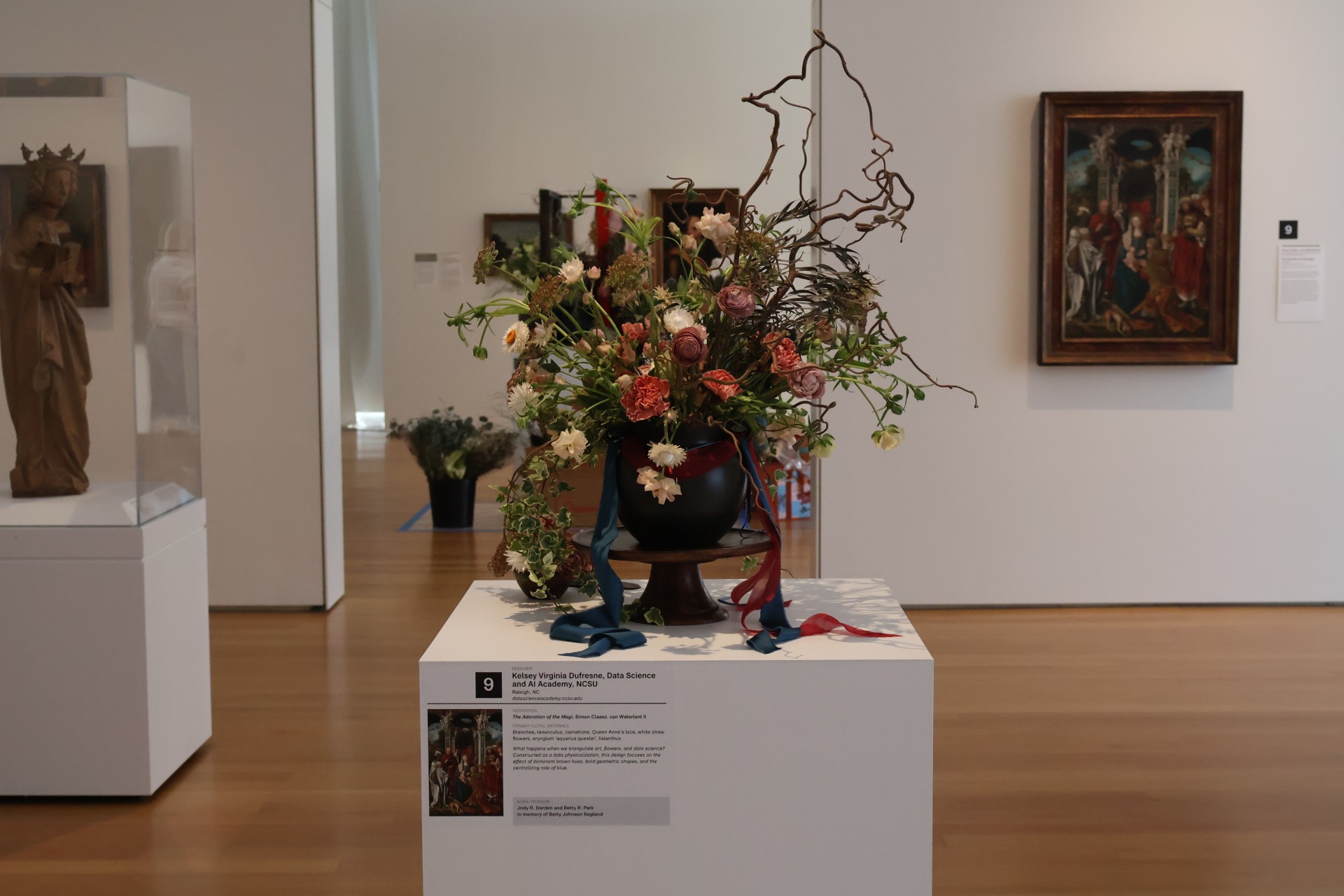 Floral arrangement displayed in a museum setting, placed on a pedestal. The arrangement includes branches and various flowers, some tied with blue and red ribbons. In the background, there are framed paintings and sculptures.