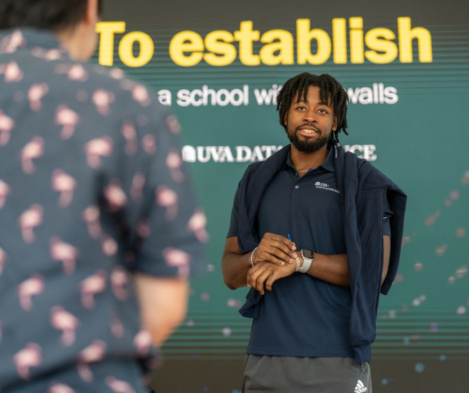 An MSDS Student Ambassador in a navy polo shirt stands in front of a large media wall that reads "To establish a school without walls" with a UVA Data Science logo underneath. Another individual, seen from the back, is facing them in conversation.