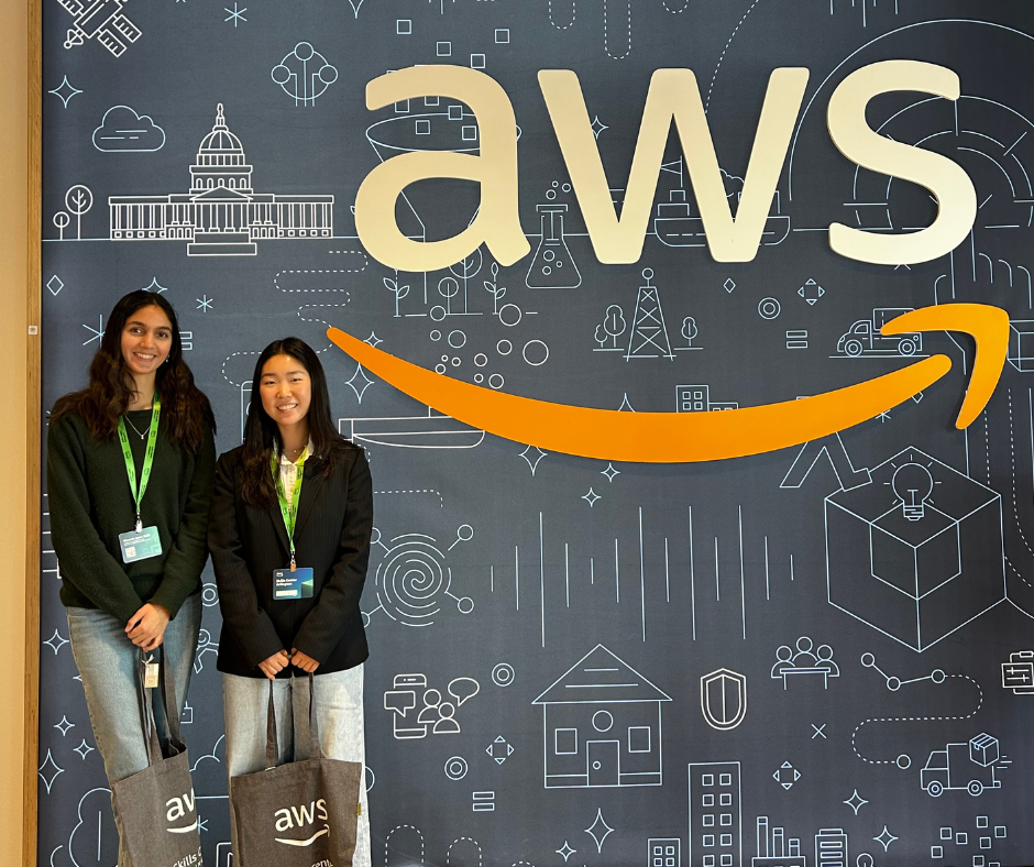 Rachel and Tara Udani stand next to the AWS logo wearing badges and carrying AWS bags.