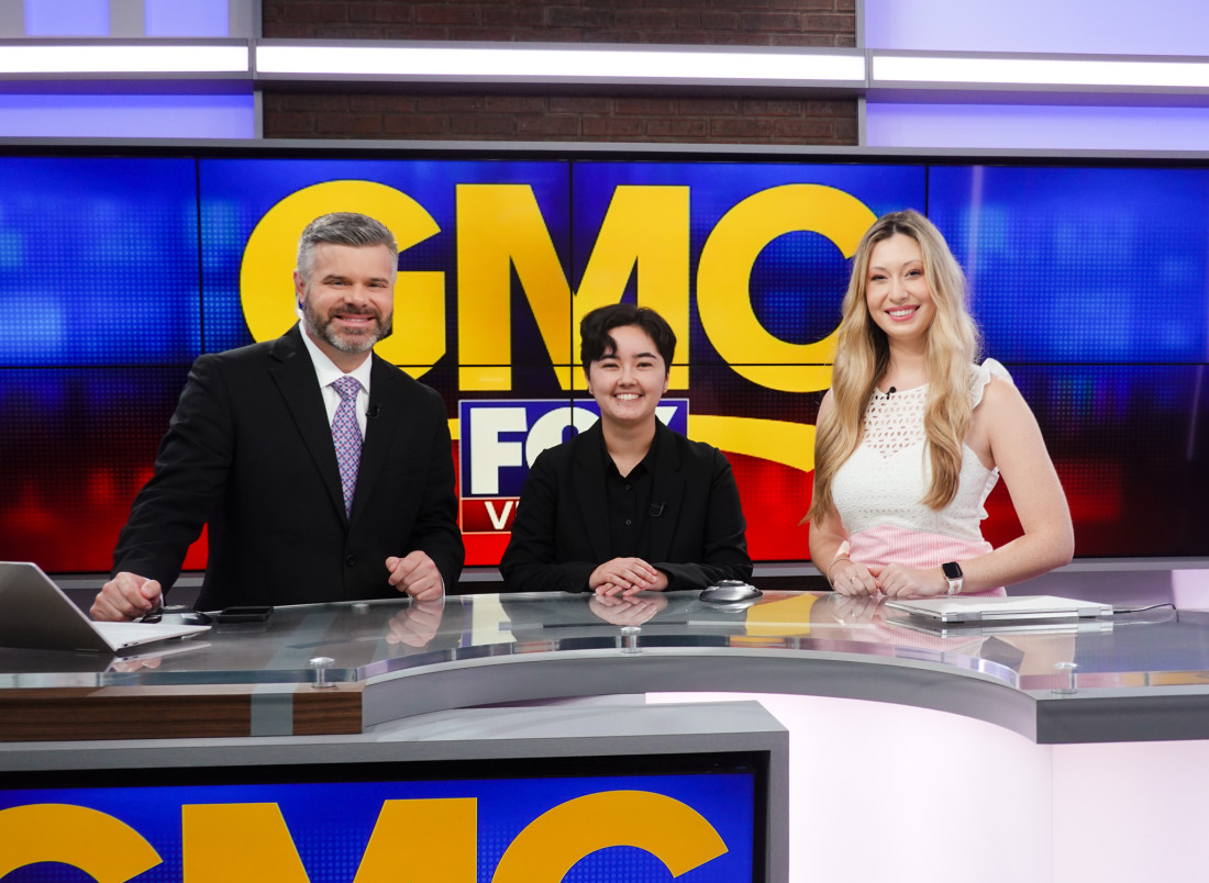 Christine poses with two news anchors in front of the GMC logo.