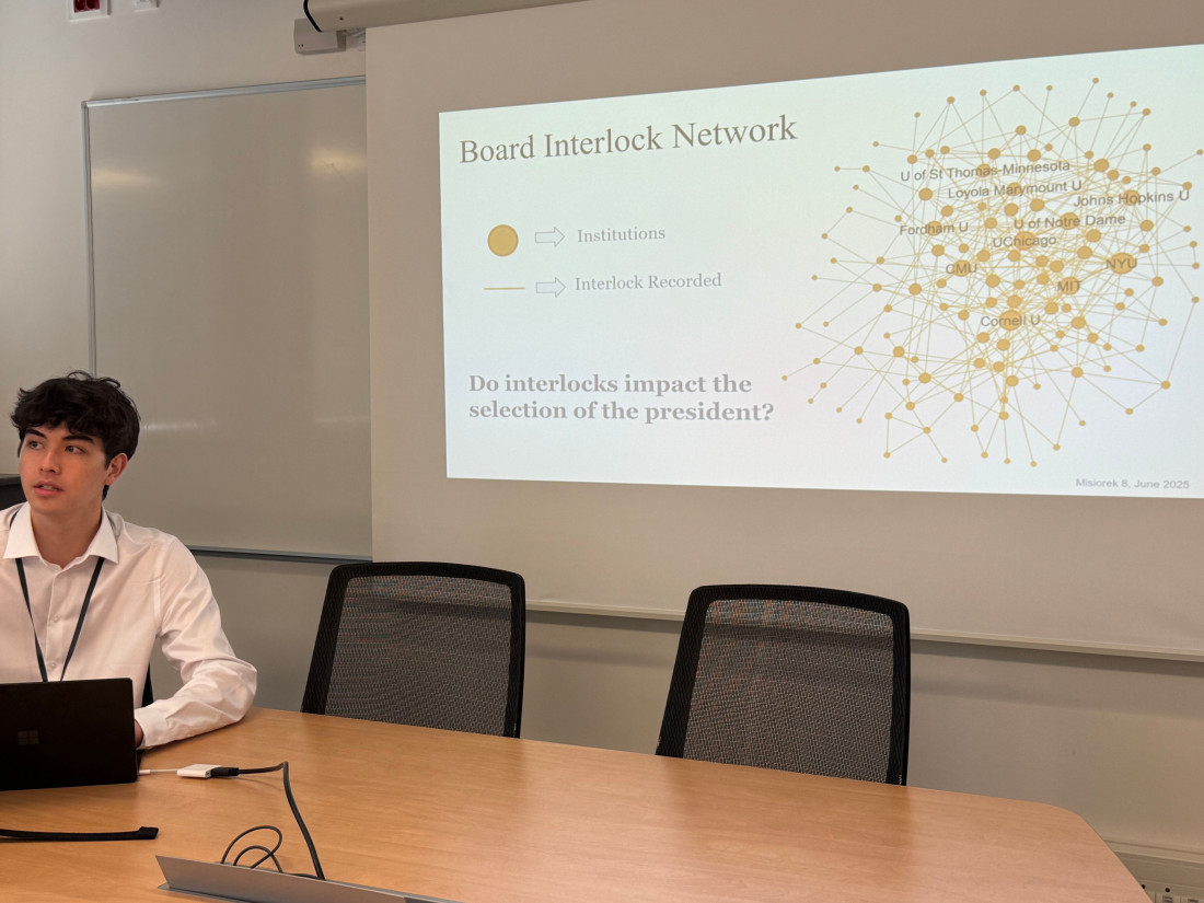 Student researcher Ty Misiorek sits at a table with a laptop, in front of a slide that reads "Board Interlock Network, Do interlocks impact the selection of the president?"