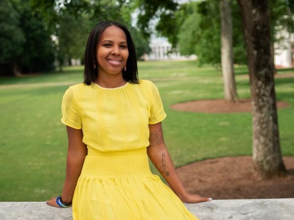 Siri Russell, Associate Dean of Diversity, Equity, and Inclusion for SDS, in a yellow dress, sits on a stone ledge in a park. She smiles confidently, surrounded by lush green grass and trees, conveying a serene, cheerful vibe.