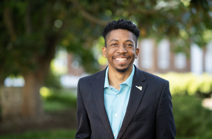 Image shows Shaka, a smiling man in a dark suit and light blue shirt stands outside. The background is a blur of trees and sunlight, conveying a cheerful, professional vibe.