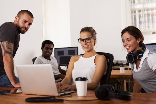 A diverse group of four young professionals collaborate happily around a laptop in a modern office. They appear focused and engaged.