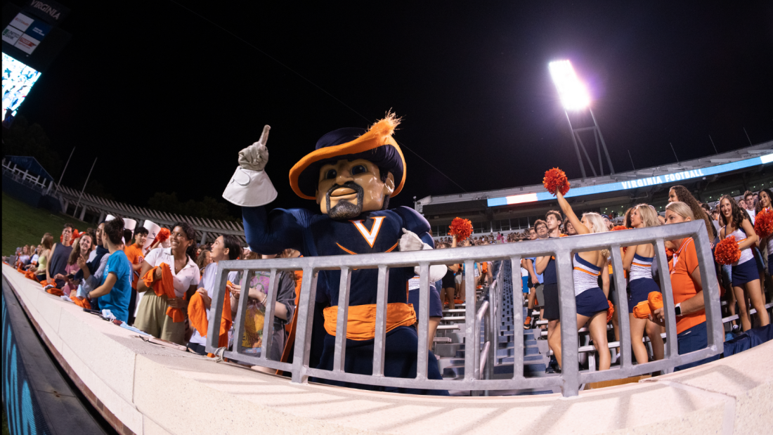 A lively football game scene at night. The UVA mascot in a large hat and cape gestures enthusiastically, surrounded by cheering fans and spirited cheerleaders.