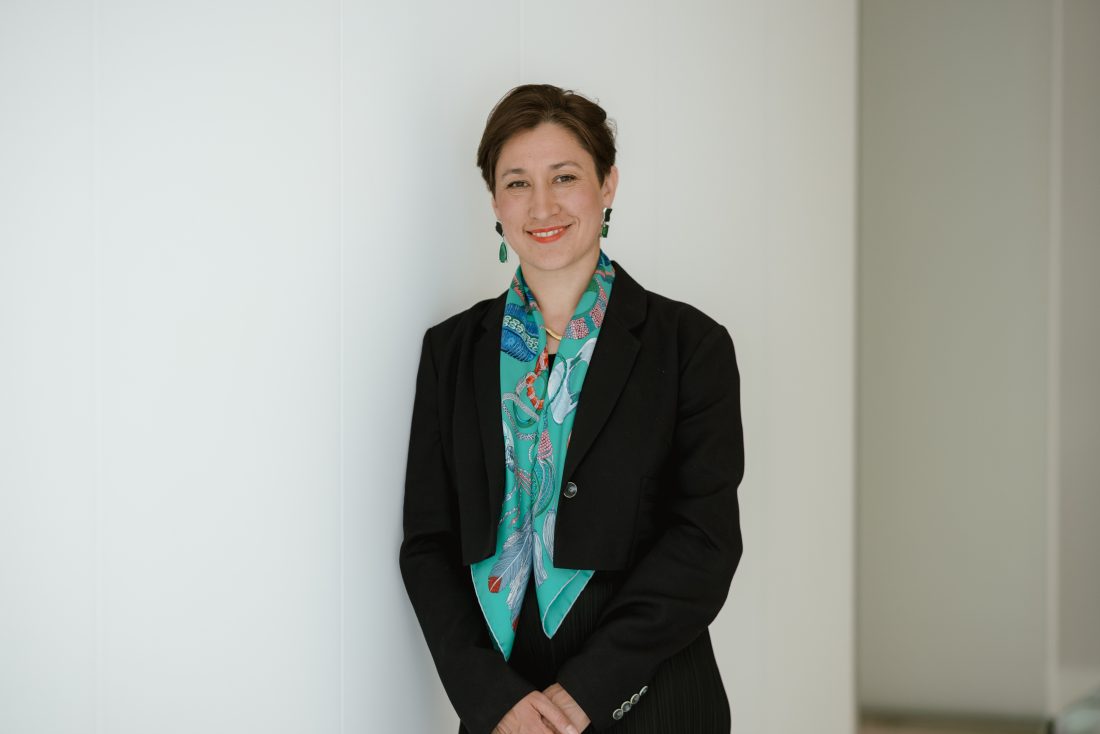 Elina Ribakova Online MSDS alumna leans against a white wall, smiling warmly. She wears a black blazer and a colorful scarf, exuding professionalism and poise.