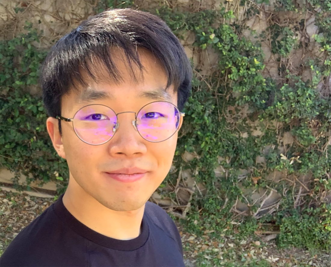 TY Kim with short black hair and glasses smiles gently at the camera. Standing outdoors against a background of ivy-covered bricks, conveying a serene and natural vibe.