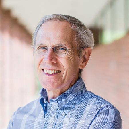 Don Brown, with short gray hair and glasses, smiles warmly. He's wearing a blue plaid shirt, and the background shows a blurred brick corridor.