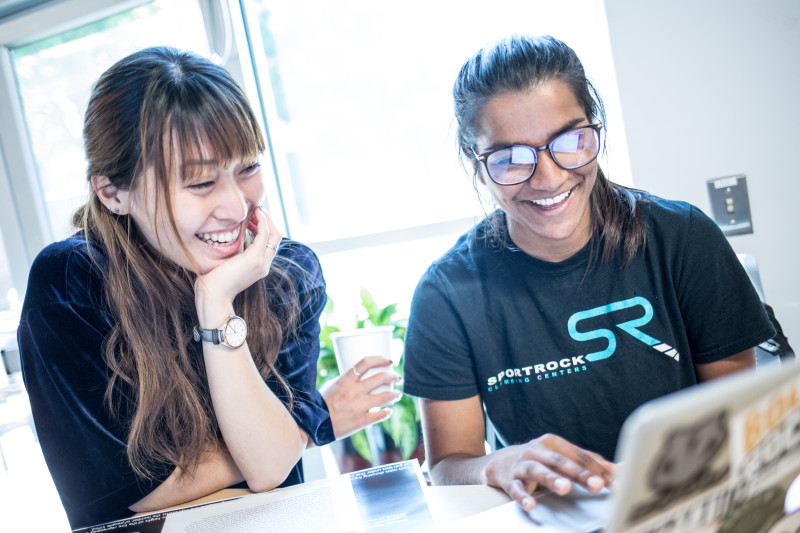Two women smiling at a laptop, one wearing glasses and a "Sportrock" t-shirt, the other holding a cup.