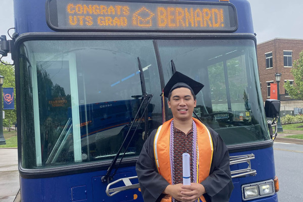 UVA Data Science student Bernard Gonzales in graduation outfit standing in front of University bus he drives