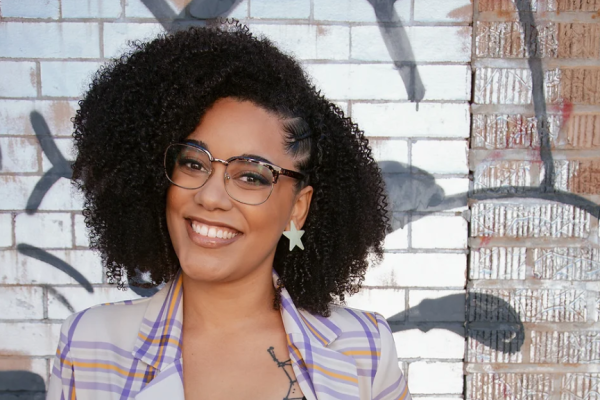 Photo of Moiya McTier standing in front of painted brick wall. She is smiling and looking at the camera. smiling and looking at the camera. 