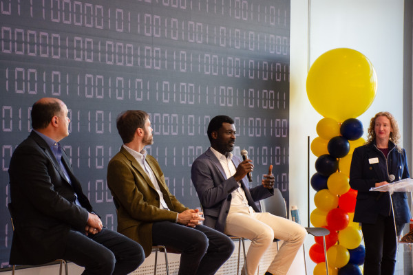 Jeffrey Blume, Alec Gosse, Prince Afriyie, and Stephanie Joynes are sitting in front of a large dark blue media wall with orange, yellow, and blue ballons off to the side. They are holding microphones ready to answer questions.