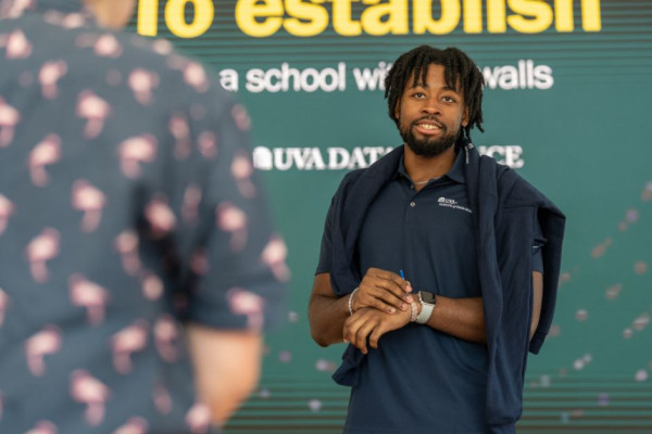 An MSDS Student Ambassador in a navy polo shirt stands in front of a large media wall that reads "To establish a school without walls" with a UVA Data Science logo underneath. Another individual, seen from the back, is facing them in conversation.