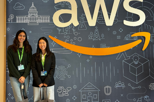 Rachel and Tara Udani stand next to the AWS logo wearing badges and carrying AWS bags.