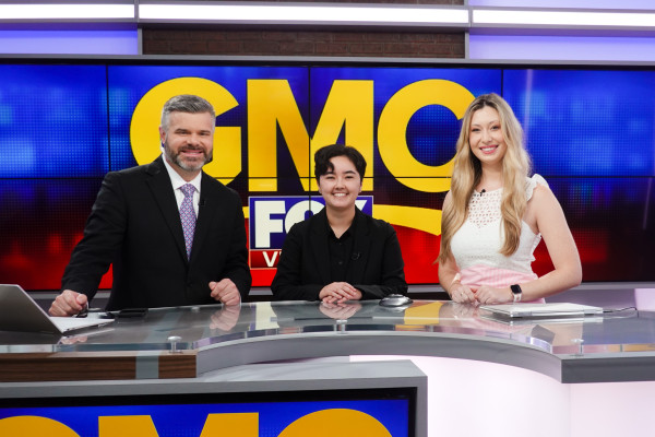 Christine poses with two news anchors in front of the GMC logo.