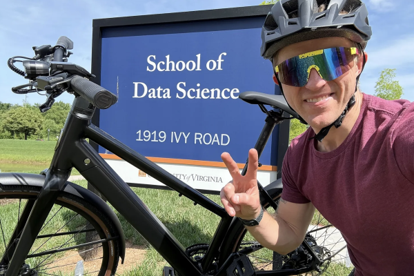 Professor Stephen Turner poses in front of the School of Data Science sign with his bike, wearing his bike helmet. 