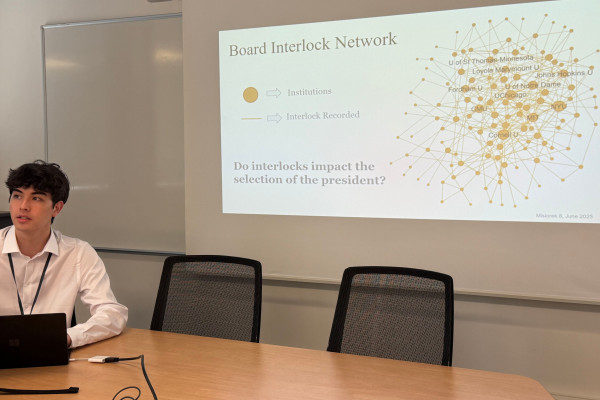 Student researcher Ty Misiorek sits at a table with a laptop, in front of a slide that reads "Board Interlock Network, Do interlocks impact the selection of the president?"