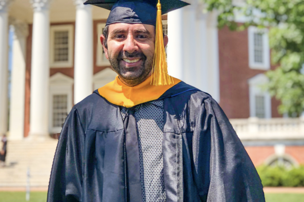 Travis wearing grad attire smiling in front of the Lawn. 