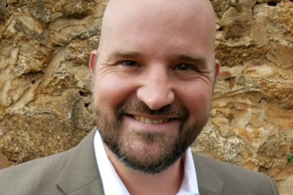 Bill Basener close up headshot wearing a tan suit. A rock-like structure is behind him. 