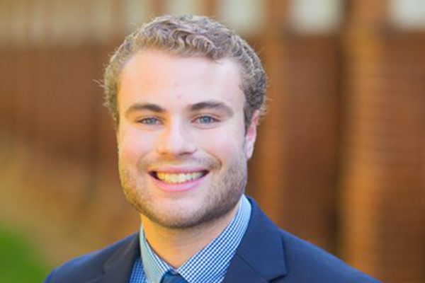 Bryan Christ with short, curly hair is pictured outdoors in front of a blurred brick background. They wear a blue checkered shirt and dark blazer.